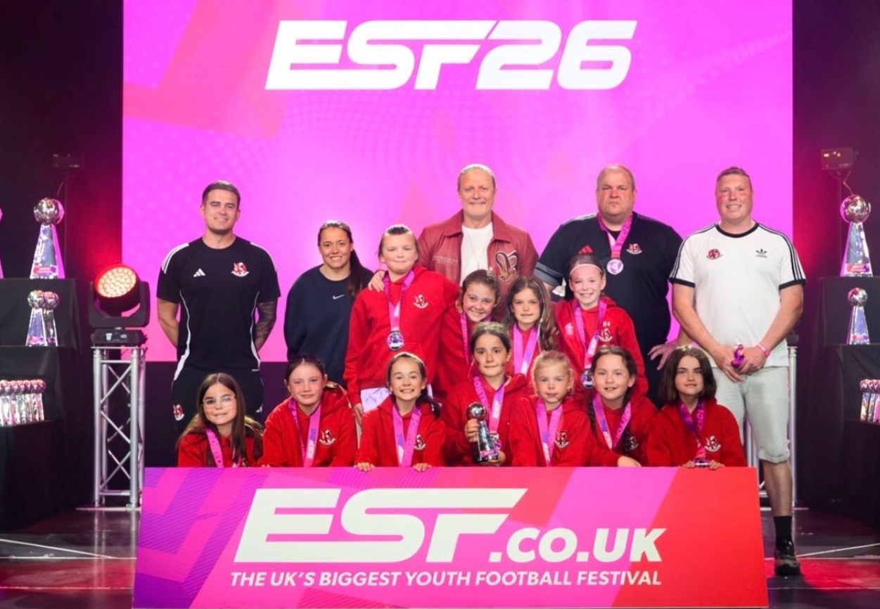 Group photo of young female football players in red jackets with medals, posing on stage at ESP26 award ceremony with coaches and trophies in the background.