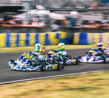 Go-kart racers speed around a curved track, colorful helmets and blue-yellow barriers in the background.