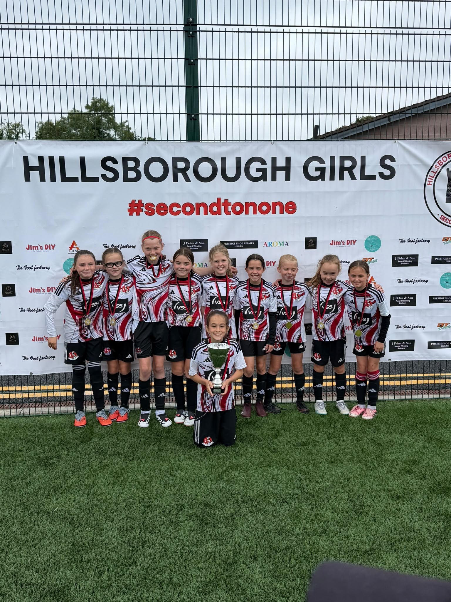 Young girls in matching soccer jerseys pose with medals and a trophy in front of a Hillsborough Girls banner.