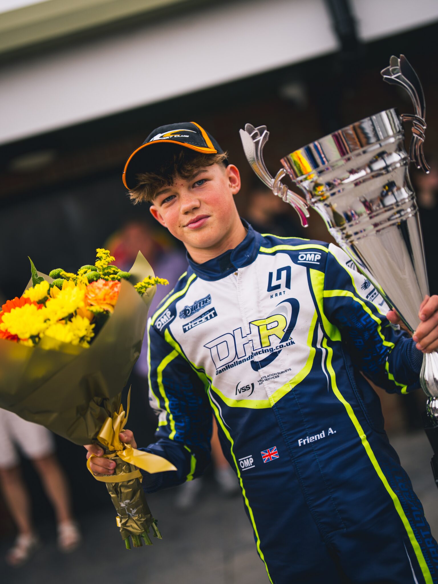 Young racing driver in blue-yellow suit holding a bouquet and a large trophy, celebrating victory.